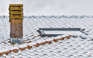 Seramik kiremitlerle kaplı karla kaplı çatı, bulutlu kış gökyüzünün altında yıpranmış baca ve tavan penceresi, sakin ve soğuk mevsimsel bir atmosfer yaratıyor..