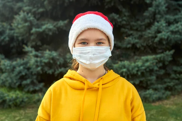 Maskeli ve Noel Baba şapkalı bir kız kameraya bakıyor. Arkaplanda ladin