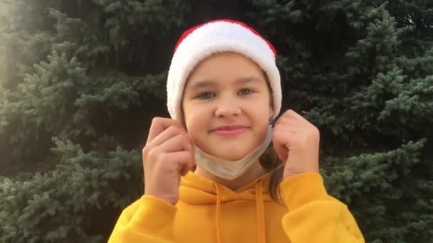 Wanita muda mengenakan topi Santa Claus mengenakan masker medis dengan latar belakang pohon Natal. Konsep merayakan Natal dengan aman selama pandemi coronavirus — Stok Video