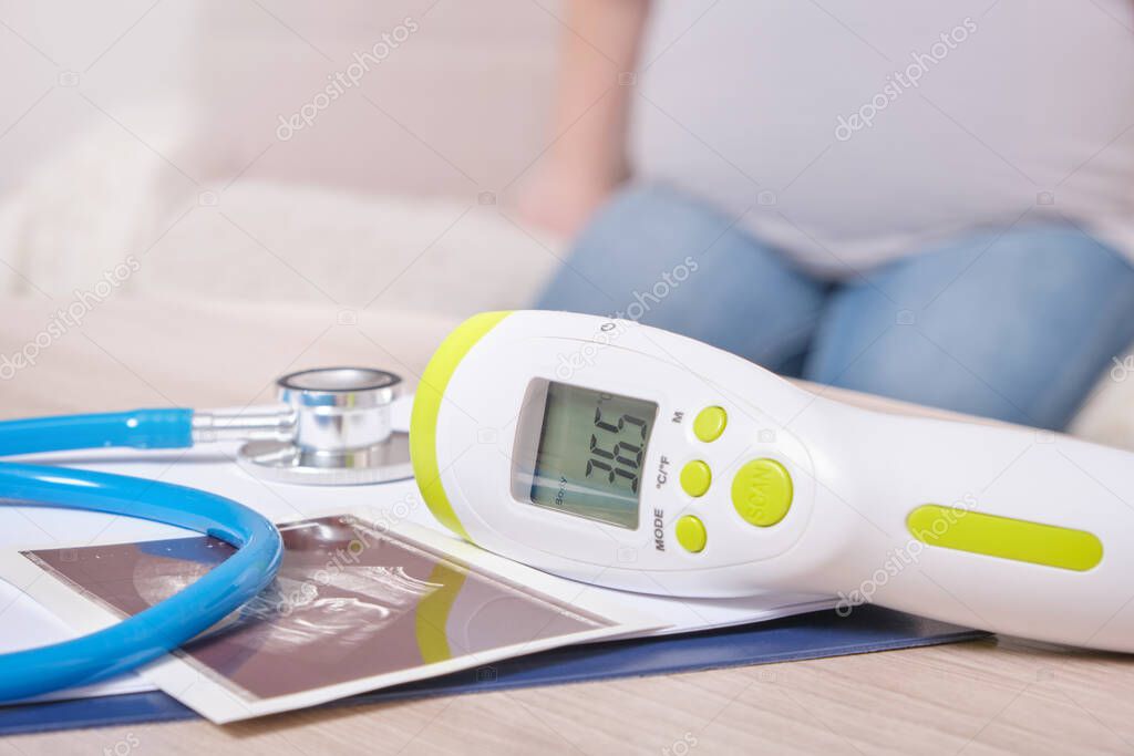 ultrasound picture, stethoscope and non-contact infrared thermometer with normal temperature on the table, pregnant woman on the background