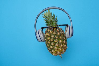 Mavi arka planda kablosuz kulaklık takan, gözlüklü, olgun bir ananas..