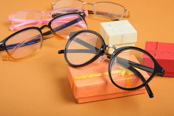 Szklanki na małym pomarańczowym pudełku, małe pudełka i różne ramki na okulary na brązowym tle przestrzeń kopia — Zdjęcie stockowe