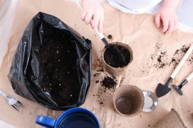 Beyaz tişörtlü bir çocuk turba tohumu kabına toprak döker, bir çocuk senen eker, bahçe aletlerini masaya koyar.