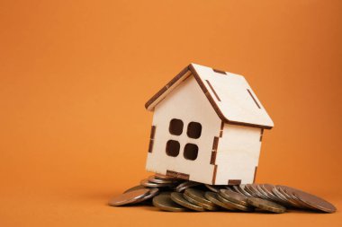 Kahverengi arka planda bir yığın madeni para üzerinde küçük ahşap ev modeli gayrimenkul konsepti satıyor, inşa ediyor veya kiralıyor