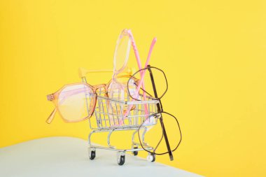 Renkli bir arka planda bir alışveriş arabasında görüşün düzeltilmesi için modaya uygun gözlük, pastel renkli kağıtlardan geometrik arkaplan, pembe bej ve açık mavi