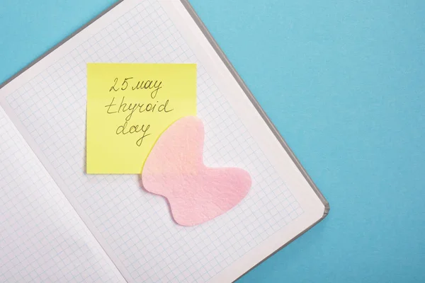 notepad, silhouette of the thyroid gland cut out of pink felt and yellow sticker on a blue background, nutrition for diseases of the thyroid gland concept copy space