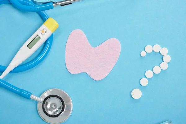 silhouette of the thyroid gland cut out of pink felt, a question mark from pills, a thermometer, ampoules and a stethoscope on a blue background, diagnostics and treatment of thyroid diseases concept