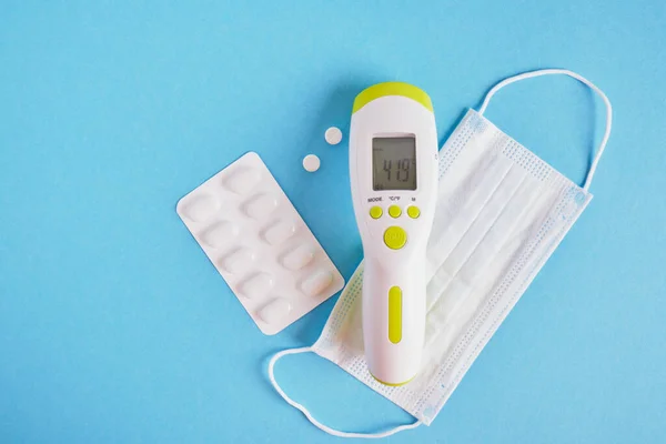 pills, face mask and electronic infrared non-contact thermometer that shows the deadly temperature on a blue background top view