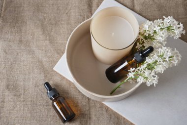 Beyaz leylaktan bir dal, bardakta aroma mumu ve doğal keten arkaplanda aroma yağı bulunan kahverengi şişeler.