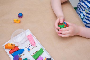 Çocukların elleri masada plastikten heykel yapıyor, bir kız plastik fotokopi aletiyle oynuyor.