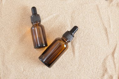 Kum arka planında damlatıcısı olan kahverengi cam şişeler, kozmetik yağ şişeleri veya logo ve etiketsiz serum, boş ve taklit kozmetik ürünü