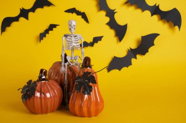 Sarı arka planda üç seramik balkabağı ve plastik iskelet, siyah kağıt yarasalardan kesilmiş, cadılar bayramı arkaplanını kopyala