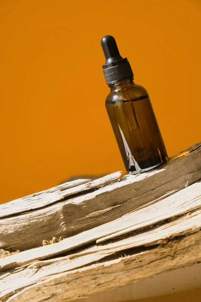 Glass cosmetic bottle with a dropper on a log on a brown background with bright sunlight. The concept of natural cosmetics, natural essential oil