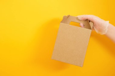Tek kullanımlık plastik bir eldiven, sarı arka planda bir karton kutu taşır. Temassız teslimat konsepti, tasarımın için boş bir paket.