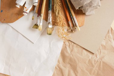 Bir dizi fırça, tüplerdeki boyalar, ahşap bir palet, kartondaki beyaz tuval ve keten arka plandaki kağıt, sanatçının atölye konsepti.