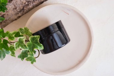 Bej arka planda seramik bir podyumda kozmetik kremi için boş siyah kavanoz, ağaç kabuğu ve yeşil yapraklar. Doğal kozmetik konsepti