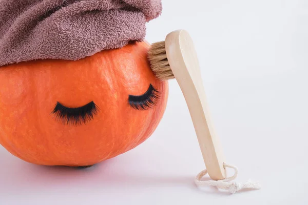 wooden face massage brush, pumpkin with false eyelashes and towel on gray background, face peeling with dry brush massage