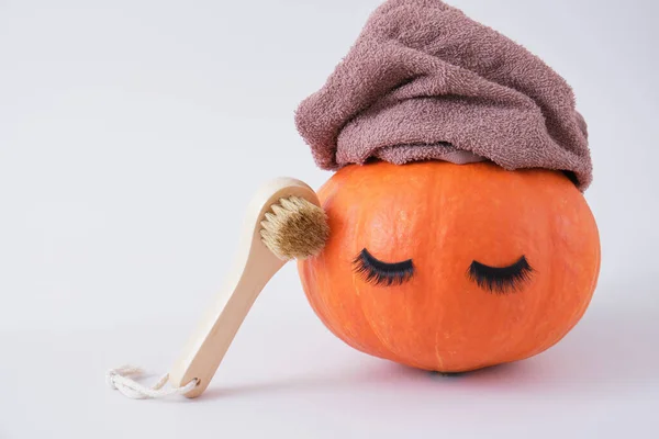 wooden face massage brush, pumpkin with false eyelashes and towel on gray background, face peeling with dry brush massage