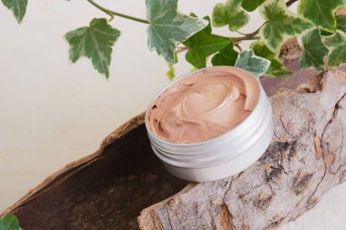 Kabuktaki alüminyum kavanozda pembe kil, açık bej arka planda yeşil yapraklı bir bitki doğal kozmetik konsepti.