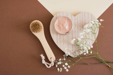 Tahta yüz masajı fırçası, boş sahte alüminyum açık kozmetik krem kavanozu pembe maske ve bej kahverengi arka planda güzel pembe ve beyaz zambak çiçekleri tahta kalp şeklinde.