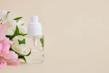 Beyaz sprey kapaklı şeffaf boş cam parfüm şişesi ve bej arka planda küçük zambak çiçekleri, kozmetik maketi
