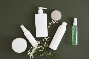 Çiçekler, farklı kozmetik kavanozlar ve koyu yeşil arka planda üç farklı beyaz şişe doğal kozmetik paketleme. Güzellik ürünleri konsepti.