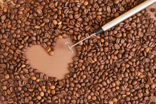 heart made of coffee beans and metal handheld milk steamer on brown background. Handheld frother or foam maker close up. copy space