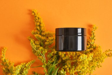 Siyah boş krema kavanozu ve turuncu arka planda sarı mimoza çiçeği. Sıfır atık çevre dostu spa ürünü. Organik cilt bakımı kozmetik.