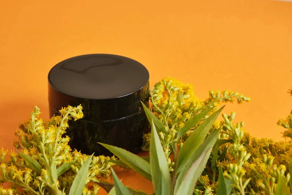 Siyah boş krema kavanozu ve turuncu arka planda sarı mimoza çiçeği. Sıfır atık çevre dostu spa ürünü. Organik cilt bakımı kozmetik.