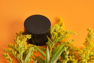 Siyah boş krema kavanozu ve turuncu arka planda sarı mimoza çiçeği. Sıfır atık çevre dostu spa ürünü. Organik cilt bakımı kozmetik.