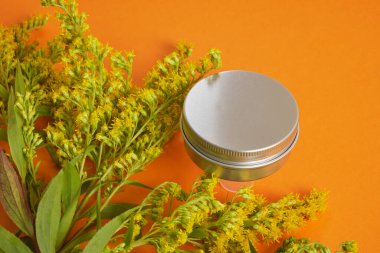 Alüminyum boş krema kavanozu ve turuncu arka planda sarı mimoza çiçeği. Sıfır atık çevre dostu spa ürünü. Organik cilt bakımı kozmetik.