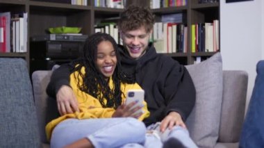 Genç çift akıllı telefondan videoya bakarken gülüyor. Erkek arkadaş ve kız arkadaş, internet ve sosyal medya için cep telefonuyla kanepede eğleniyorlar. Mutlu ortaklar ve evde teknoloji