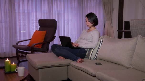 Ασιατικές γυναίκα με φορητό υπολογιστή τηλέφωνο τηλέφωνο στον καναπέ — Αρχείο Βίντεο