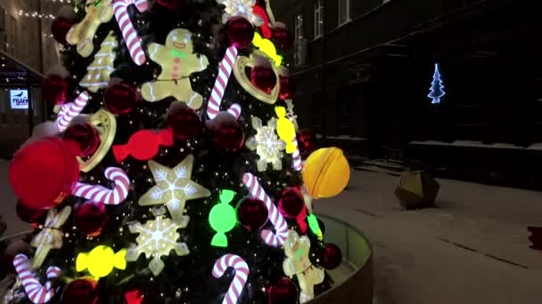 Kar yağışı sırasında karlı caddede süslenmiş Noel ağacı. Noel oyuncaklarıyla Noel ağacı süslemesi, şeker kamışlı karamel, Noel topları ve zencefilli kurabiye adam. Aydınlatılmış Noel ağacı oyuncakları — Stok video