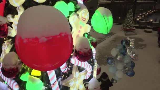 Geschmückter Weihnachtsbaum auf verschneiter Straße bei Schneefall. Christbaumschmuck mit Spielzeug, Zuckerrohrkaramellen, Kugeln und Lebkuchenmann. Beleuchtetes Neujahrsbaumspielzeug — Stockvideo