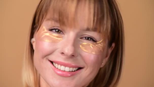 Beautiful smiling woman with short hair in gold eye patches looking in camera and smiling over colorful gold background. Beauty concept — Stock Video