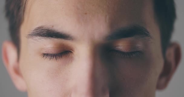 Vista de cerca de un joven tranquilo está abriendo los ojos y mirando a la cámara en la pared de fondo gris en el estudio — Vídeos de Stock