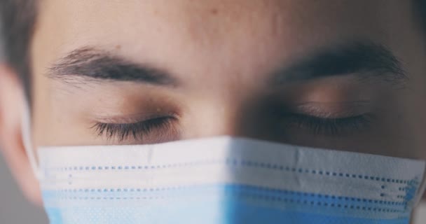 Mladý muž v lékařské masce otevírá oči a dívá se přímo do kamery. Detailní portrét ve studiu. Virus concept. — Stock video