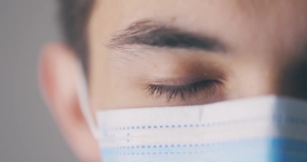 Mladý muž v lékařské masce otevírá oči a dívá se přímo do kamery. Detailní portrét ve studiu. Polovina obličeje. Virus concept. — Stock video