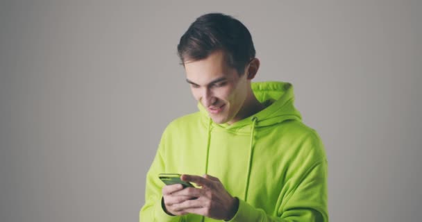 Jovem bonito em roupas verdes usando mensagem de leitura de telefone inteligente sorrindo, rindo. Retrato filmado em estúdio em fundo cinza — Vídeo de Stock