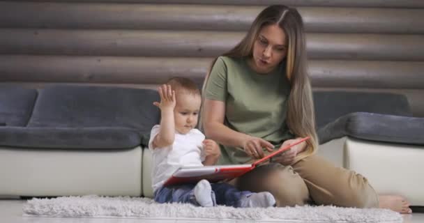 Młoda matka i jej synek bawią się książką siedząc na podłodze w domu. Mama bawić się z małym chłopcem w domu. — Wideo stockowe