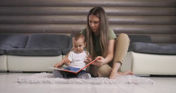 Mladá matka čte knížku svému malému synovi, který zvědavě naslouchá, zatímco sedí doma na podlaze. Matka hrát s malým chlapečkem doma.