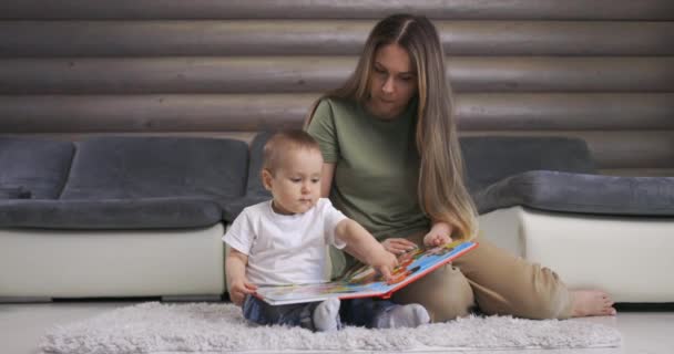 Ung mor og hendes lille søn leger med bogen, mens de sidder på gulvet derhjemme. Mor leger med lille baby dreng hjemme. – Stock-video