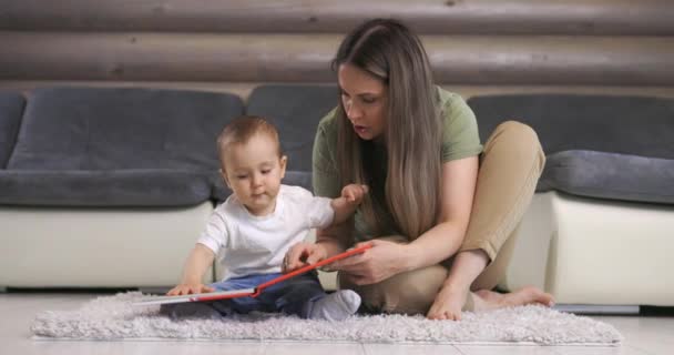 Молодая мать и ее маленький сын играют с книгой, сидя на полу дома. Мать играет с маленьким мальчиком дома. — стоковое видео