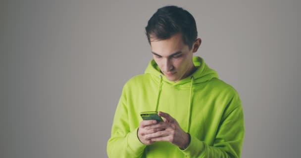 Homem bonito jovem em uma camisola brilhante usando smartphone no fundo do estúdio cinza. Usando a tecnologia moderna - aplicativos, redes sociais. — Vídeo de Stock