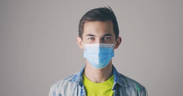 Un caucazian chipeş cu mască medicală de protecţie arată degetele mari, ca un gest. Două mâini. Un tip fericit pe fundal gri. Câştigător. Succes. Modelul masculin pozitiv zâmbește camerei . — Videoclip de stoc