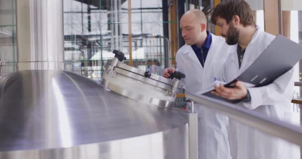 Twee blanke mannen die in een brouwerij werken. Een mannelijke brouwer controleert de metingen met een tablet. — Stockvideo