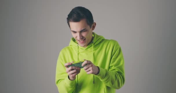 Jovem bonito em suor verde jogando jogo no smartphone na parede do estúdio cinza. Usando a tecnologia moderna - aplicativos, redes sociais. — Vídeo de Stock