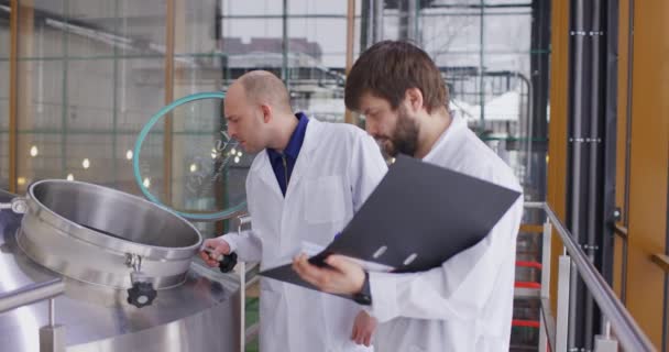 Twee blanke mannen die in een brouwerij werken. Een mannelijke brouwer controleert de metingen met een tablet. — Stockvideo