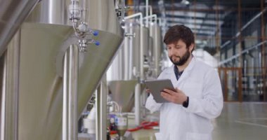 Beyaz laboratuvar önlüklü bir adam ekipman göstergesine bakıyor ve tablete veri girişi yapıyor. Erkek bira fabrikası işçisi paslanmaz bira fıçılarının önünde duruyor.